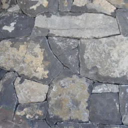 Fragment ściany zewnętrznej wykończonej nieregularnymi kamieniami w odcieniach szarości i beżu, z widoczną rynną i fragmentem okna w tle.