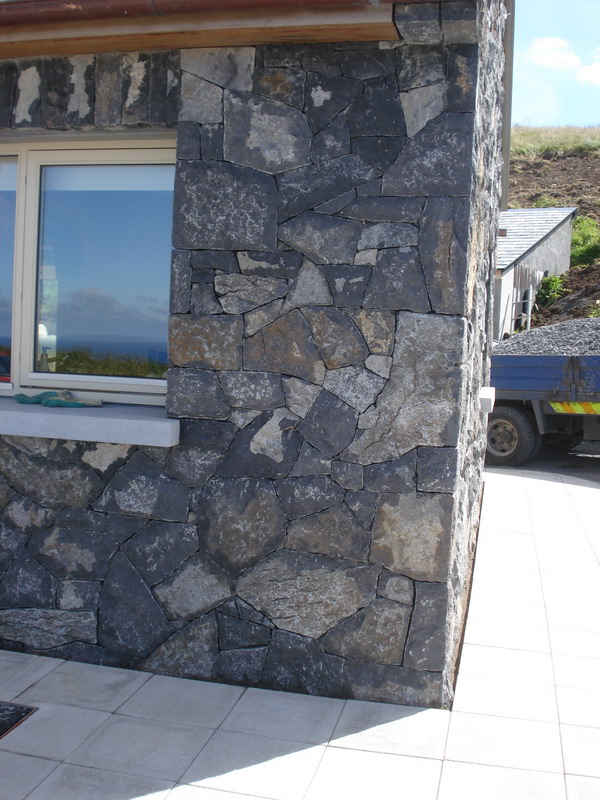 Ściana budynku wykończona nieregularnymi kamieniami w odcieniach szarości i czerni, widoczne okno z odbijającym się niebem i morzem, obok zaparkowana ciężarówka z gruzem.