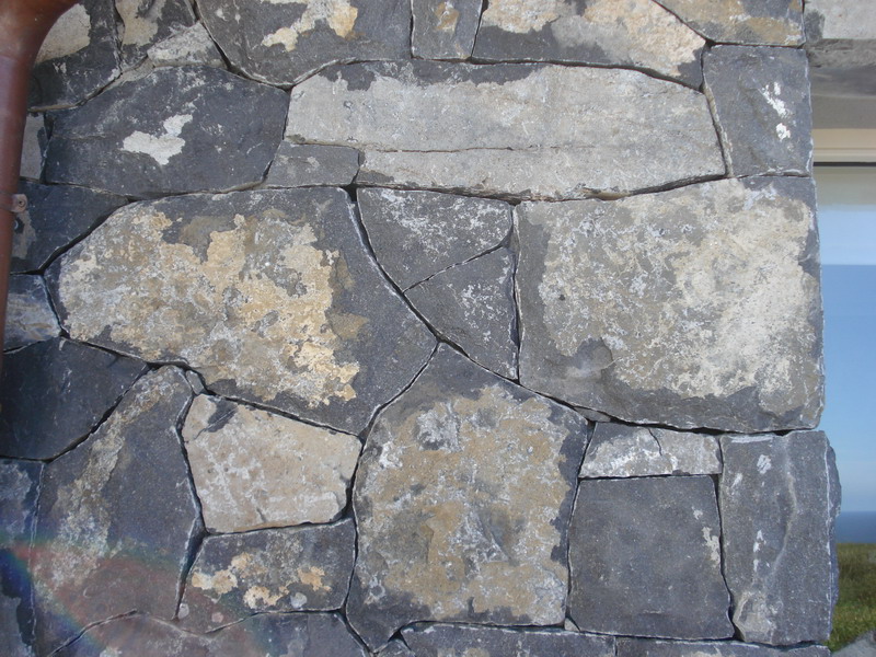 Fragment ściany zewnętrznej wykończonej nieregularnymi kamieniami w odcieniach szarości i beżu, z widoczną rynną i fragmentem okna w tle.