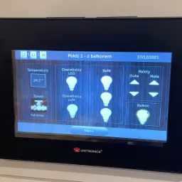 Panel sterowania inteligentnego domu z ekranem dotykowym, pokazujący ustawienia temperatury, oświetlenia LED i sufitowego, rolet oraz balkonu, z logo Unitronics w dolnej części.