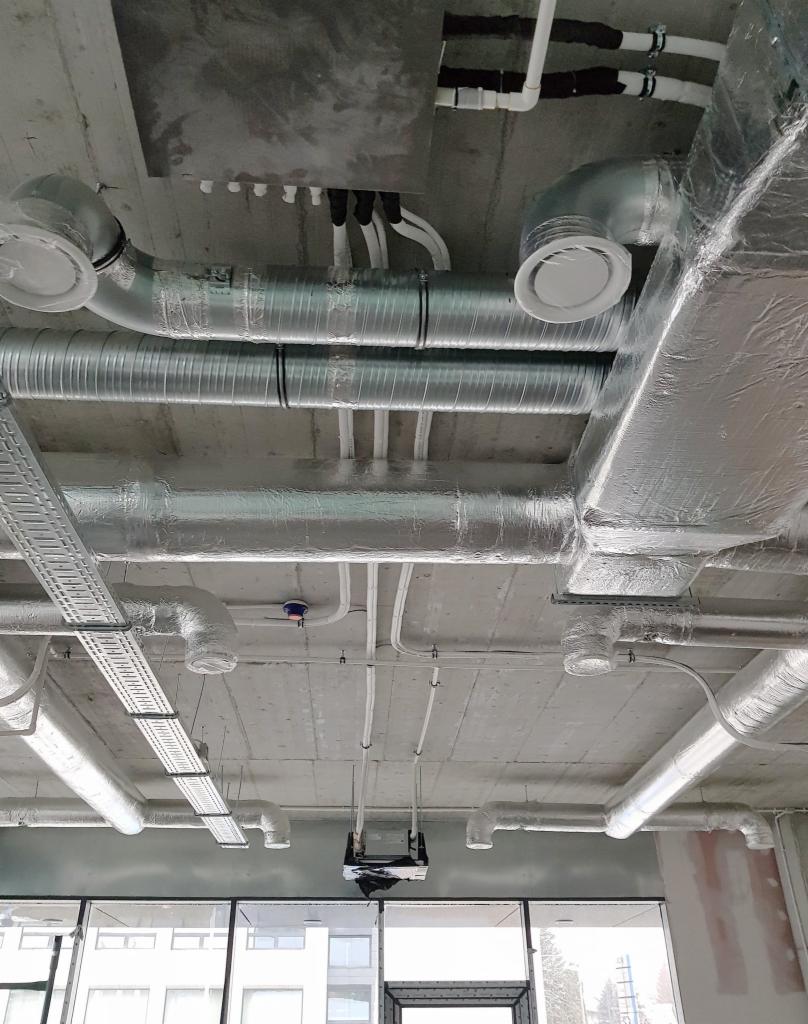 Instalacja wentylacyjna i klimatyzacyjna pod betonowym sufitem w surowym stanie, widoczne rury aluminiowe, kanały wentylacyjne, kable i kratki wentylacyjne, częściowo zabezpieczone folią, w tle...