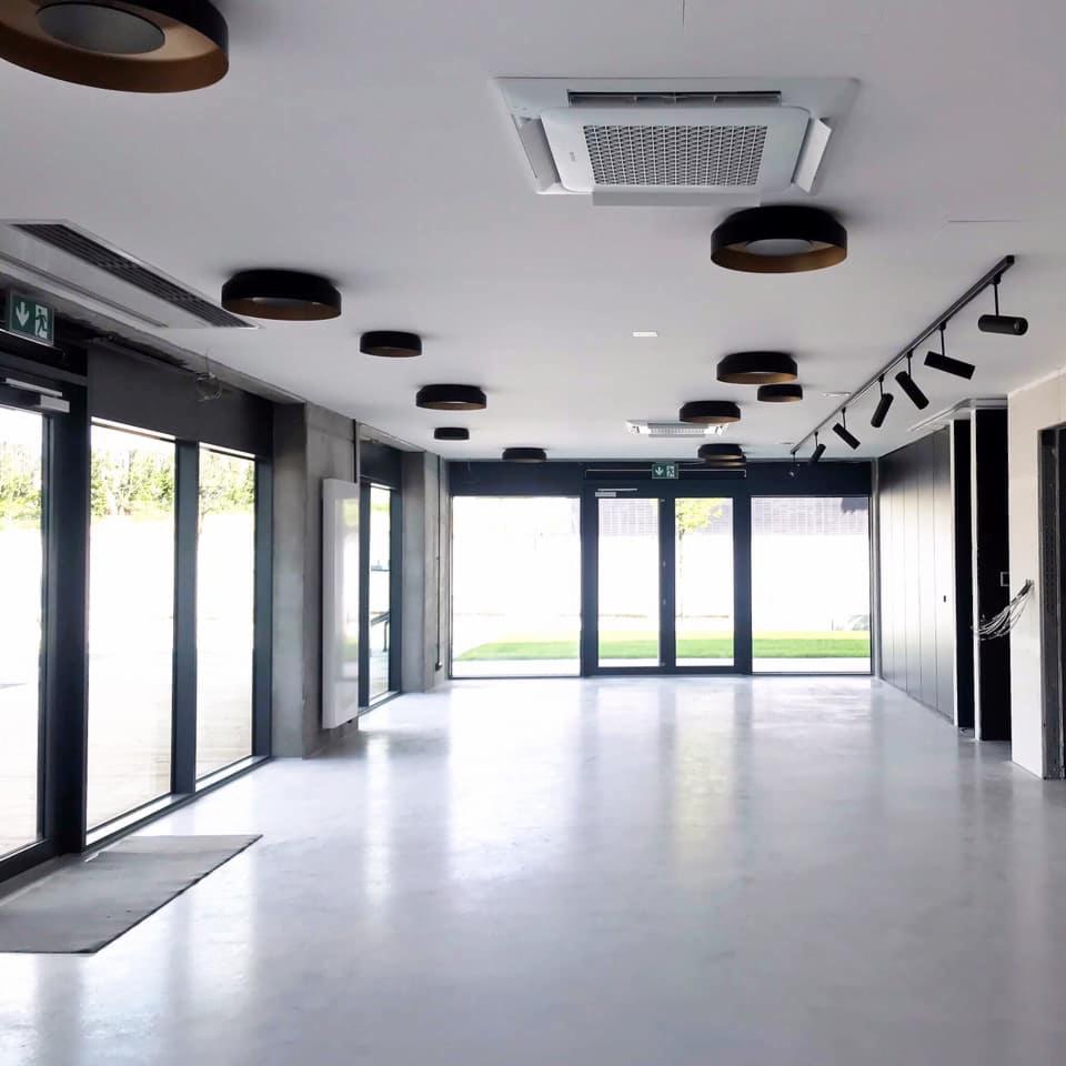 Wnętrze biurowe z zamontowaną kasetonową jednostką klimatyzacyjną w suficie podwieszanym, widoczne także okrągłe lampy sufitowe i system oświetlenia szynowego.