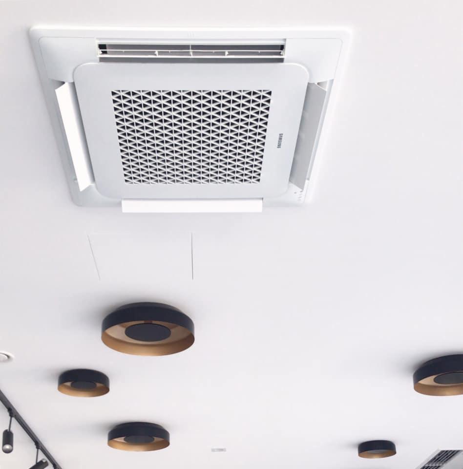 Kasetonowa jednostka wewnętrzna klimatyzacji Samsung zamontowana na białym suficie z widocznymi okrągłymi lampami sufitowymi w kolorze czarnym ze złotym wnętrzem.