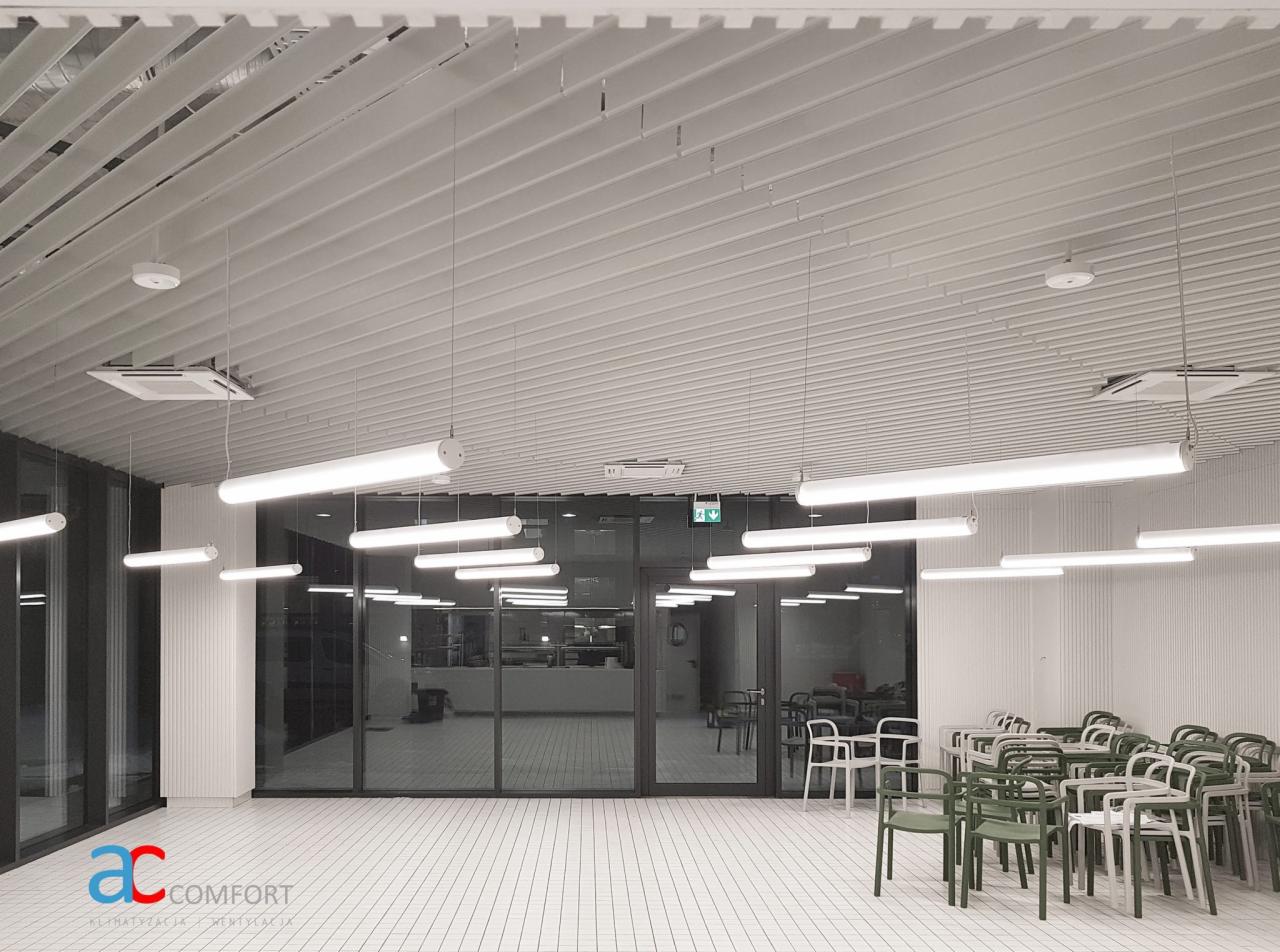 Wnętrze biurowe z systemem wentylacji pod sufitem, widoczne lampy i kratki wentylacyjne, biała podłoga z płytek, krzesła w tle, logo firmy w lewym dolnym rogu.