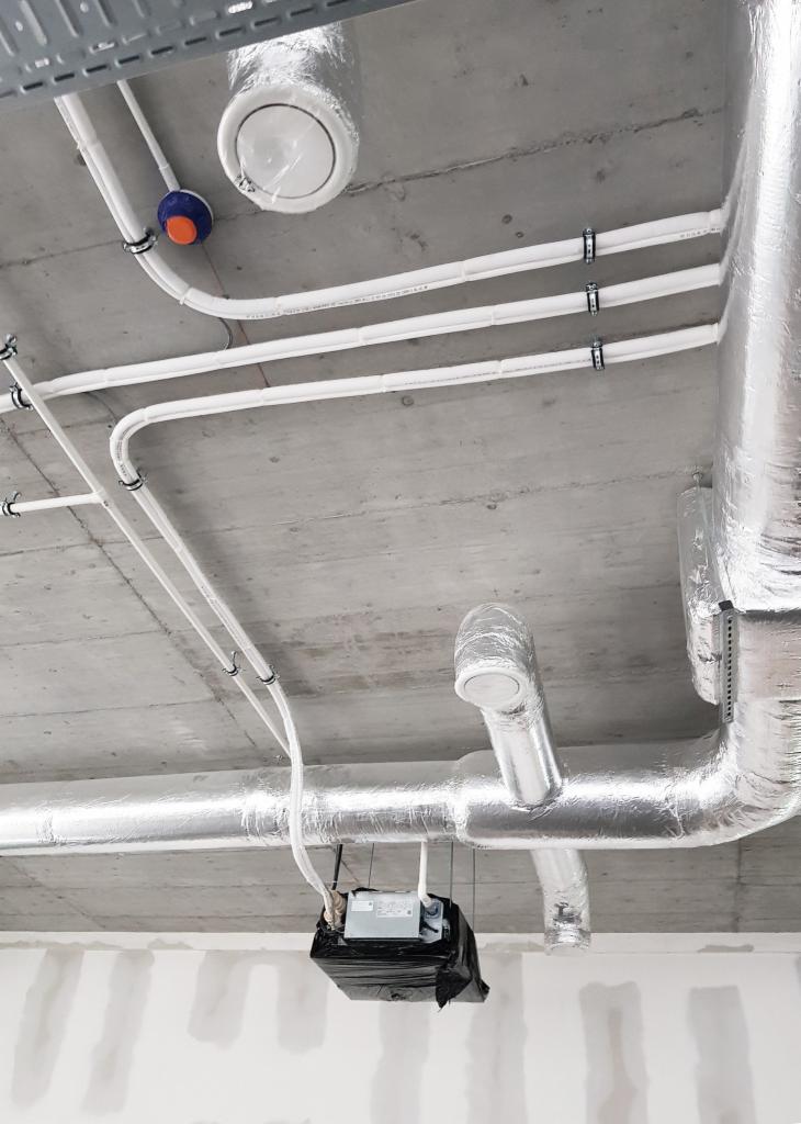 Widok na sufit z instalacją wentylacyjną: rury w izolacji aluminiowej, białe rury PCV z uchwytami, okrągły dyfuzor i jednostka centralna w czarnym worku ochronnym, surowy beton sufitu.