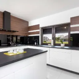 Nowoczesna kuchnia z białymi szafkami, czarnym blatem i okładziną ścienną z motywem drewna, z widocznym zestawem do herbaty na blacie i oknem z roletami.