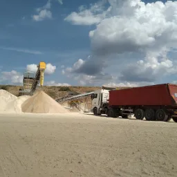Ciężarówka z czerwoną naczepą obok hałd piasku i taśmociągu na tle błękitnego nieba z białymi chmurami.