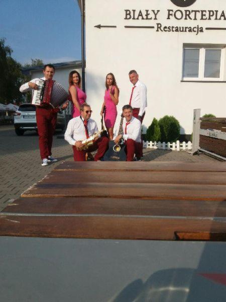 Sześcioosobowy zespół muzyczny w bordowych spodniach i różowych sukienkach pozuje przed budynkiem restauracji 'Biały Fortepian', mężczyzna z akordeonem, saksofonista, gitarzysta i dwie wokalistki.