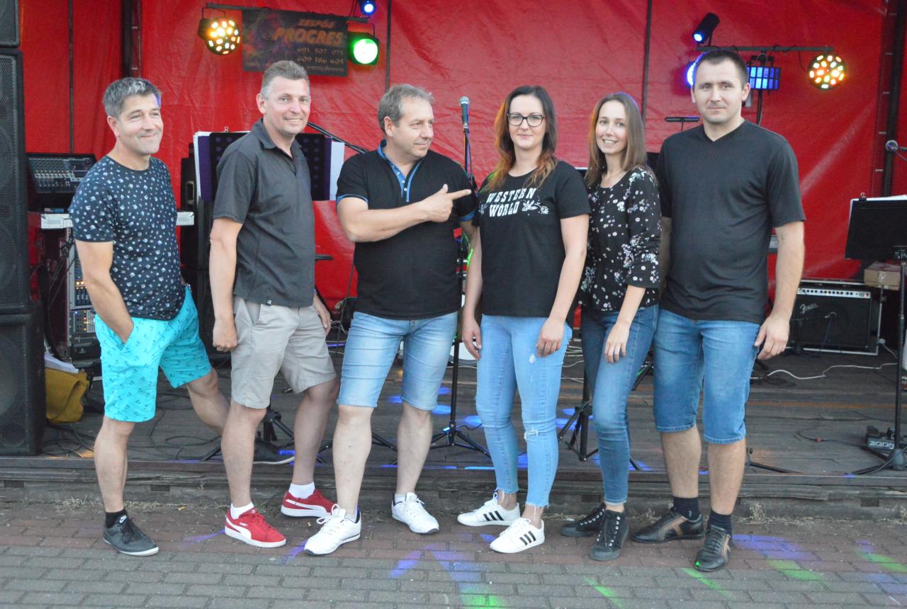 Zespół muzyczny 'Progres' w składzie sześcioosobowym pozuje na scenie z czerwonym tłem i reflektorami, jeden z członków wskazuje palcem na koleżankę z zespołu.