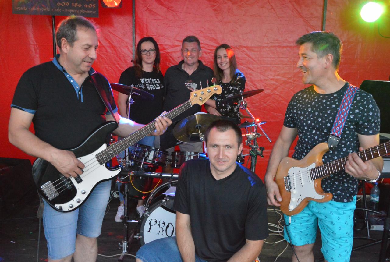 Zespół muzyczny w trakcie występu: basista gra na czarnej gitarze basowej, gitarzysta na jasnej gitarze elektrycznej, perkusja z logo 'PRO' w tle, oświetlenie sceniczne.