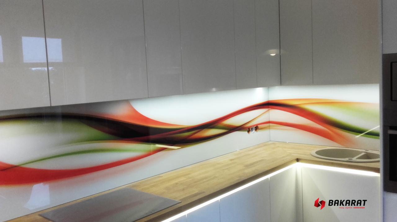 Szklany panel kuchenny z abstrakcyjnym motywem fal w kolorach czerwieni, zieleni i czerni, zamontowany między blatem a szafkami, podświetlony od dołu.