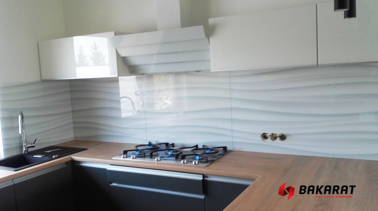 Szklana ściana nad blatem kuchennym z motywem fal, odbijająca światło, uzupełniona o białe szafki wiszące, czarny zlew i czteropalnikową płytę gazową.