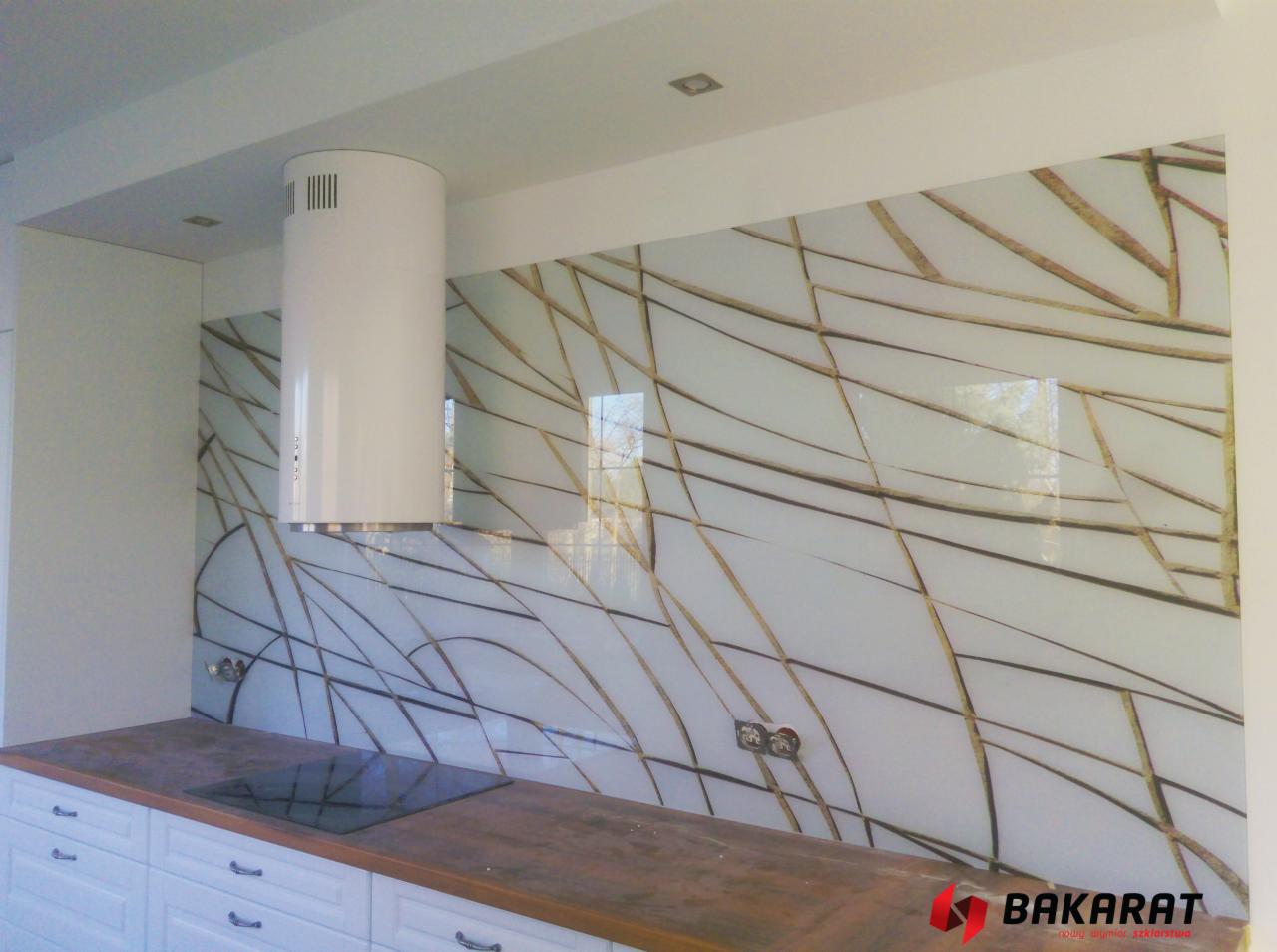 Szklany panel ścienny w kuchni z abstrakcyjnym wzorem w odcieniach bieli i złota, widoczny okap kuchenny cylindryczny i fragment blatu z płytą indukcyjną.