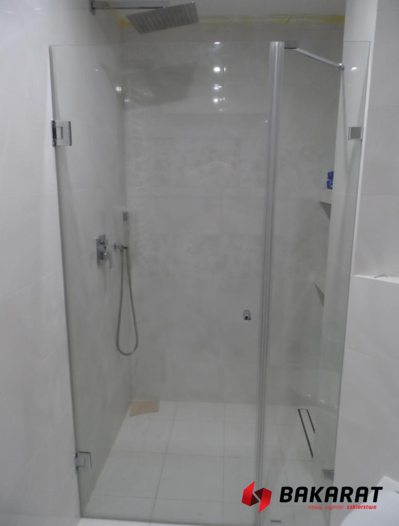 Szklana kabina prysznicowa z chromowanymi zawiasami i deszczownicą, widok z zewnątrz, białe płytki na ścianach i podłodze, logo firmy Baccarat w rogu.