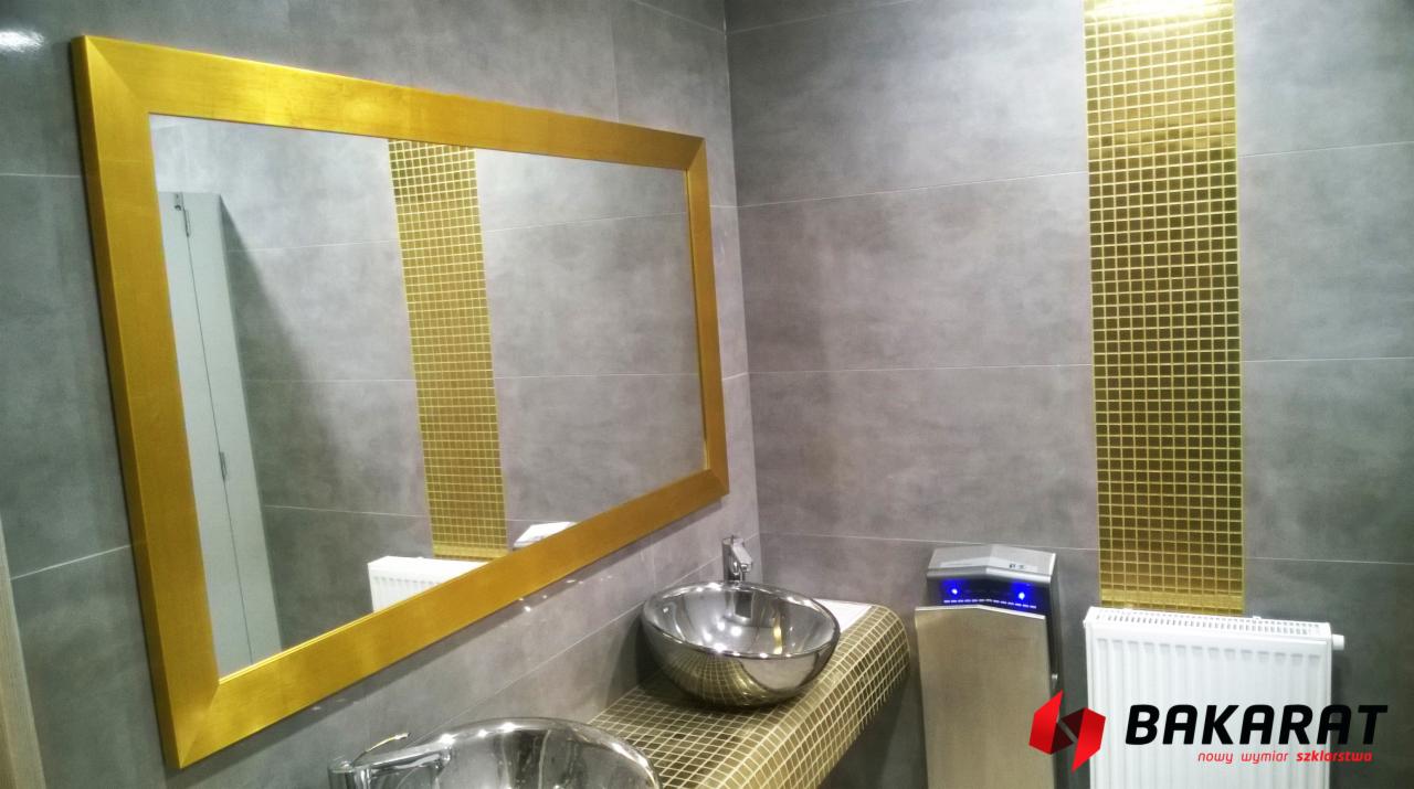 Łazienka z dużym lustrem w złotej ramie, srebrnymi umywalkami i pasem złotej mozaiki na szarej ścianie z płytek, widoczny grzejnik i suszarka do rąk, logo firmy szklarskiej w prawym dolnym rogu.