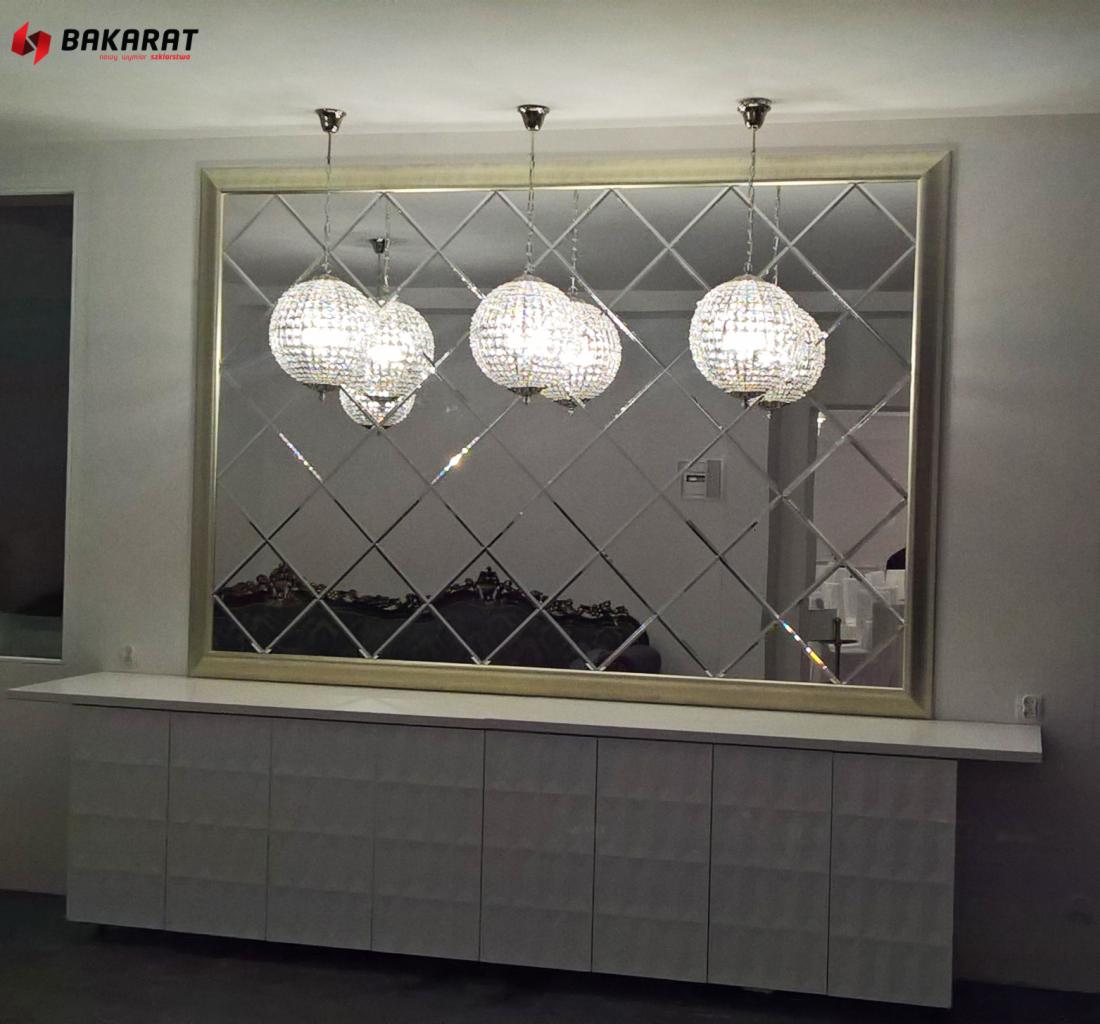 Dekoracyjne lustro ścienne z fazowanych rombów, oprawione w złotą ramę, zawieszone nad białą komodą, odbijające kryształowe żyrandole.