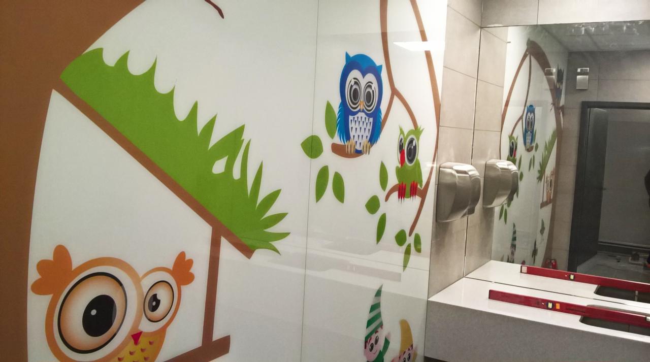 Szklana ściana w łazience z motywem bajkowych sów i krasnali, odbijająca się w lustrze nad umywalką, gdzie widać poziomice; obok zamontowane suszarki do rąk
