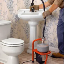 Hydraulik w rękawicach używa elektrycznej spirali do przepychania zatkanej umywalki na tle ściany w kwiaty