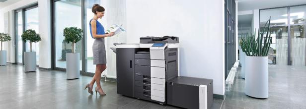 Kobieta przegląda dokumenty obok nowej, wielofunkcyjnej kserokopiarki A3 w nowoczesnym biurze z roślinami doniczkowymi.