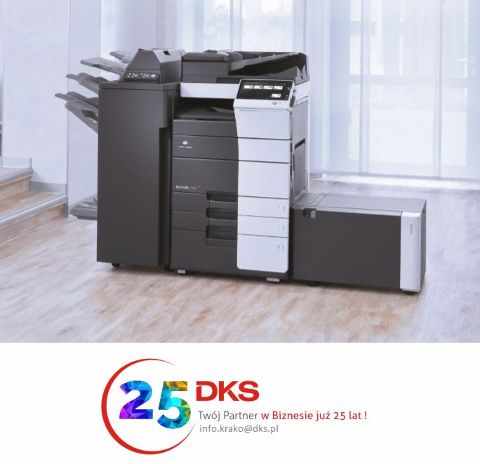 Nowoczesna, czarno-biała kserokopiarka wielkoformatowa z dodatkowym modułem podajnika na tle jasnego biura, widoczny fragment schodów i logo firmy w dolnej części kadru.