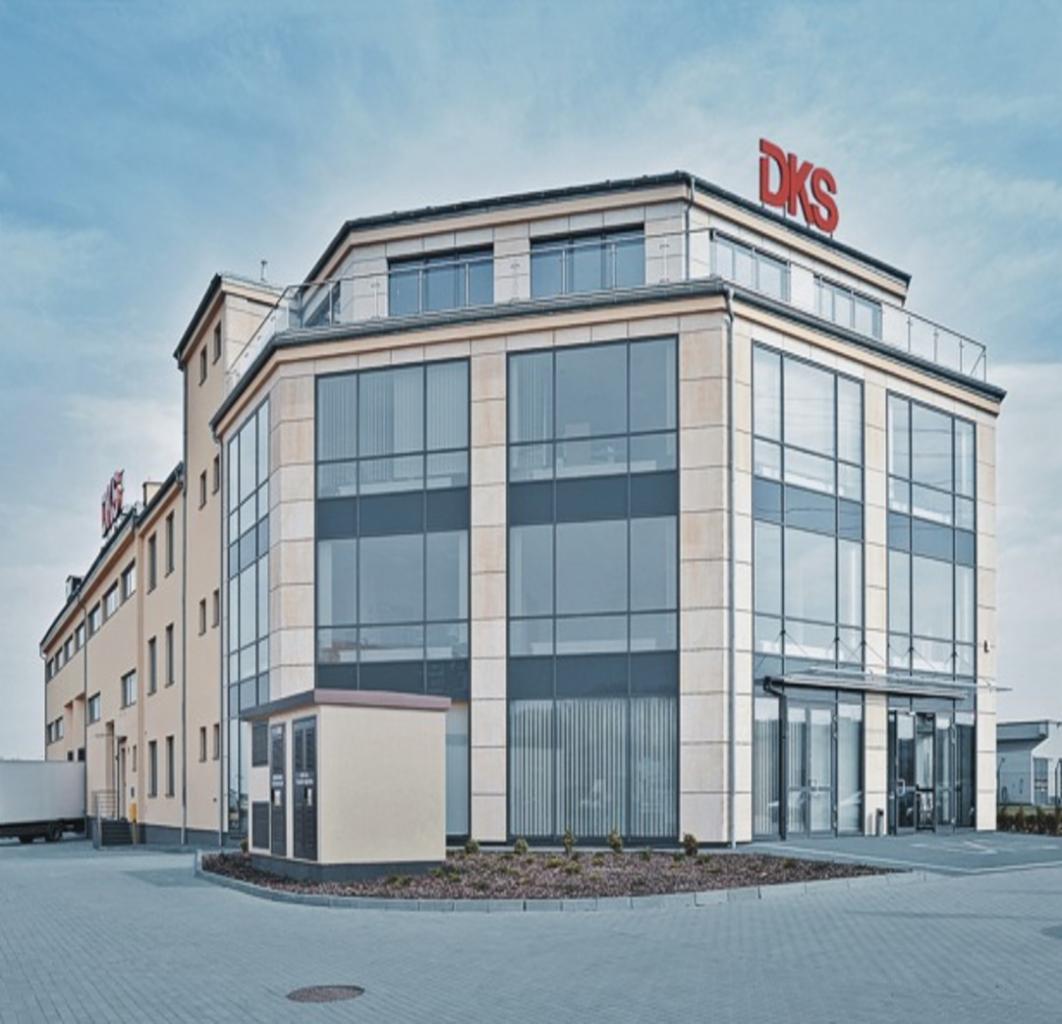 Nowoczesny budynek biurowy z dużymi oknami i logo DKS na dachu, widoczny fragment parkingu i przyczepy ciężarówki.