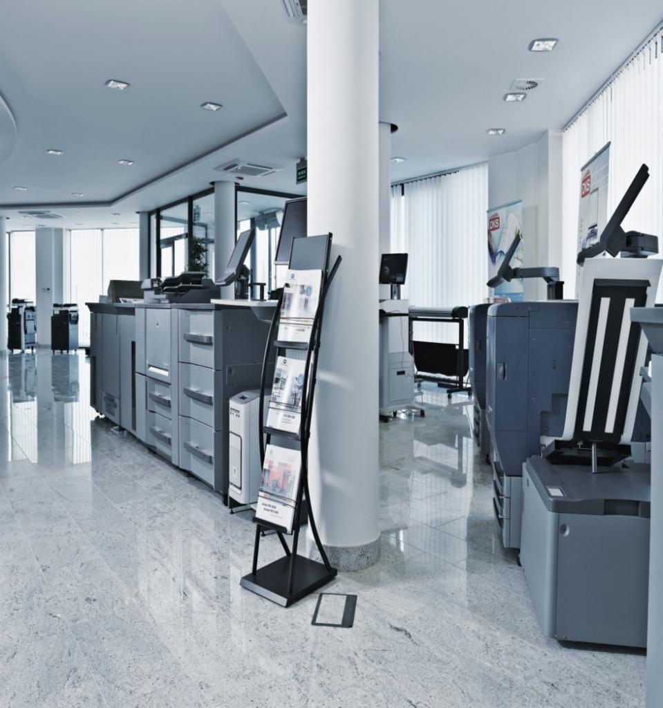 Przestronne biuro z rzędem wielkoformatowych kserokopiarek i stojakiem na ulotki, widok na marmurową podłogę i kolumnę.