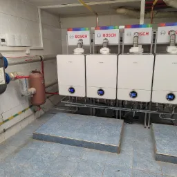 Cztery białe, wiszące piece gazowe Bosch z kolorowymi tabliczkami znamionowymi, podłączone do instalacji, widoczne elementy instalacji grzewczej i wodnej, na pierwszym planie podest z płytek.