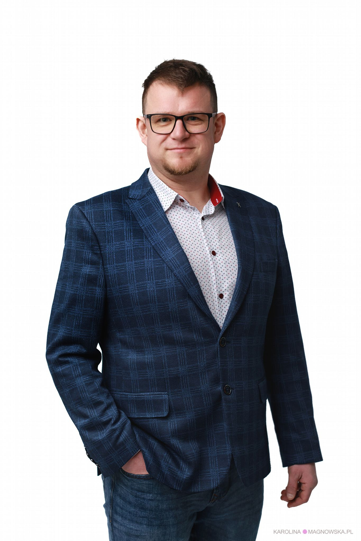 Mężczyzna w okularach i marynarce w kratę, z ręką w kieszeni dżinsów, na białym tle. Uśmiechnięty, profesjonalny portret.