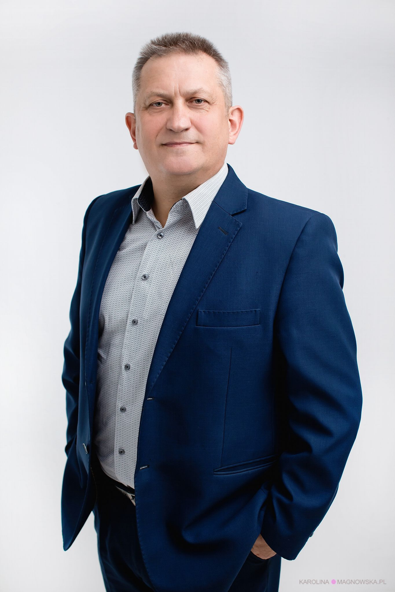 Portret mężczyzny w niebieskiej marynarce i wzorzystej koszuli na białym tle, z rękami w kieszeniach. Profesjonalny wygląd, neutralny wyraz twarzy.