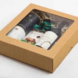 Kartonowe pudełko prezentowe z okienkiem, zawierające kawę, herbatę, butelkę wina i czekoladę, wypełnione dekoracyjnym siankiem i przewiązane zieloną wstążką.