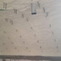 Poddasze z oknem dachowym pokryte jasną pianą izolacyjną, z widocznymi metalowymi mocowaniami i fragmentem komina wentylacyjnego.