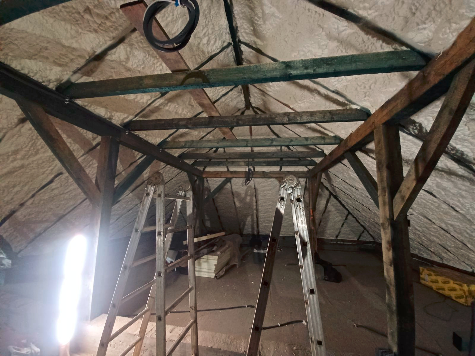 Poddasze z drewnianą konstrukcją pokryte szarą pianką izolacyjną, widoczne dwie drabiny aluminiowe i wiązka kabli pod sufitem.