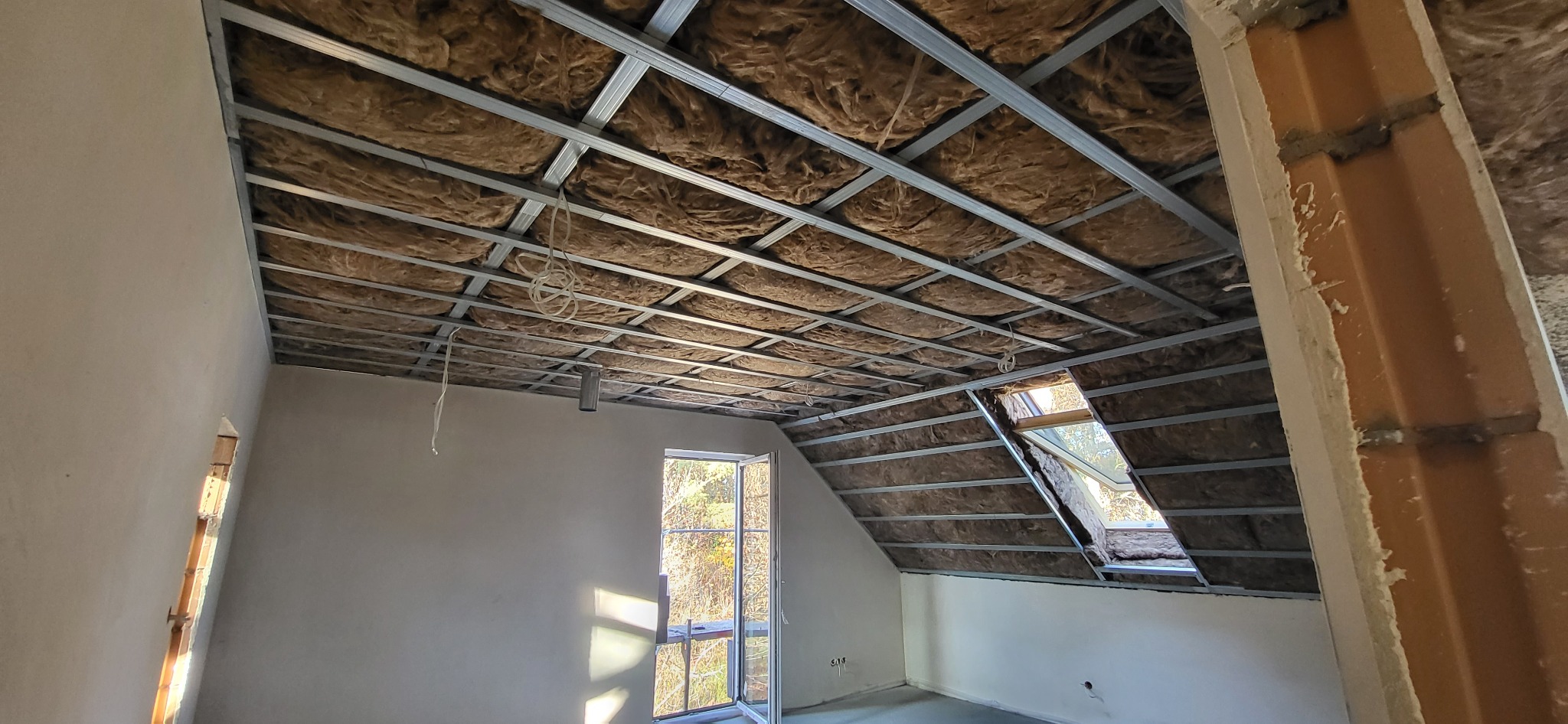 Stelaż metalowy z izolacją z wełny mineralnej poddasza, widoczne okno dachowe i niezakończone ściany z cegły.