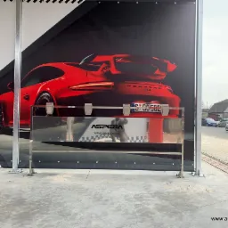 Uchwyty ze stali nierdzewnej na dywaniki samochodowe na myjni bezdotykowej, zamontowane na tle reklamy czerwonego Porsche GT3, widok z zewnątrz na chodnik i zaparkowane samochody.