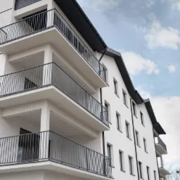 Nowoczesny budynek wielorodzinny z balkonami zabezpieczonymi metalowymi balustradami w kolorze grafitowym, widok z dołu na tle błękitnego nieba.