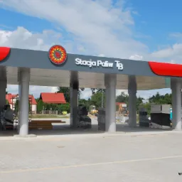 Zmodernizowana stacja paliw z szarym zadaszeniem, czerwonym obramowaniem i logo 'TB'. W tle widoczne budynki i sprzęt budowlany, sugerujące trwające prace wykończeniowe.