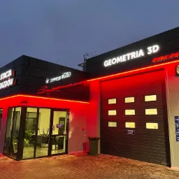 Budynek Okręgowej Stacji Kontroli Pojazdów z podświetlanym czerwonym neonem, widoczne logo 'Geometria 3D Brezelia' nad bramą wjazdową i oznakowanie reklamowe na fasadzie.