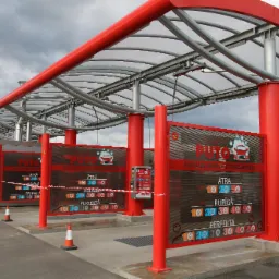 Zewnętrzna konstrukcja samoobsługowej myjni samochodowej z czerwonymi słupami i stalową konstrukcją dachu, widoczne cenniki z różnymi opcjami mycia.