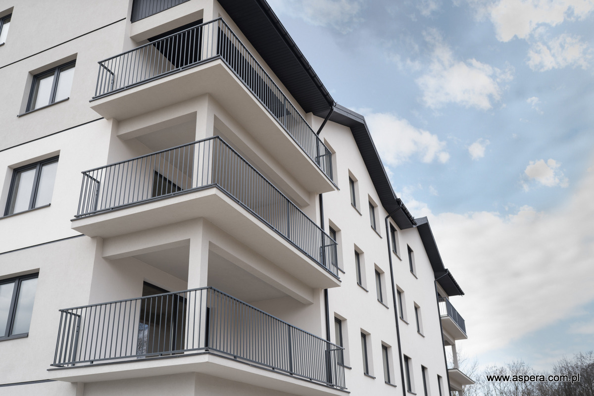 Nowoczesny budynek wielorodzinny z balkonami zabezpieczonymi metalowymi balustradami w kolorze grafitowym, widok z dołu na tle błękitnego nieba.