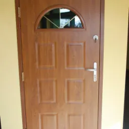 Brązowe drzwi zewnętrzne z półokrągłym przeszkleniem i srebrną klamką, zamontowane w żółtej ścianie.