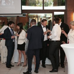 Tomasz Krzyczkowski - Grupa elegancko ubranych gości wznosi toast szampanem w sali bankietowej z projekcją w tle, oświetlenie punktowe, nowoczesny wystrój.