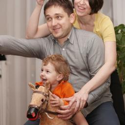 Tomasz Krzyczkowski - Rodzina: uśmiechnięty chłopiec w pomarańczowej bluzie siedzi na pluszowym koniku, wspierany przez ojca w szarej koszuli; matka zza pleców męża unosi rękę z obrączką, wszyscy patrzą w lewo