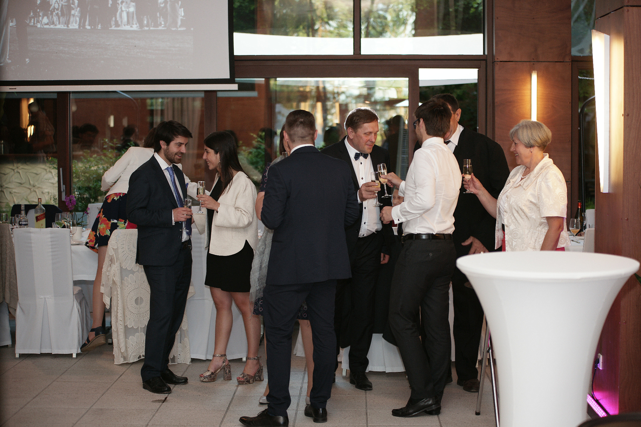 Grupa elegancko ubranych gości wznosi toast szampanem w sali bankietowej z projekcją w tle, oświetlenie punktowe, nowoczesny wystrój.