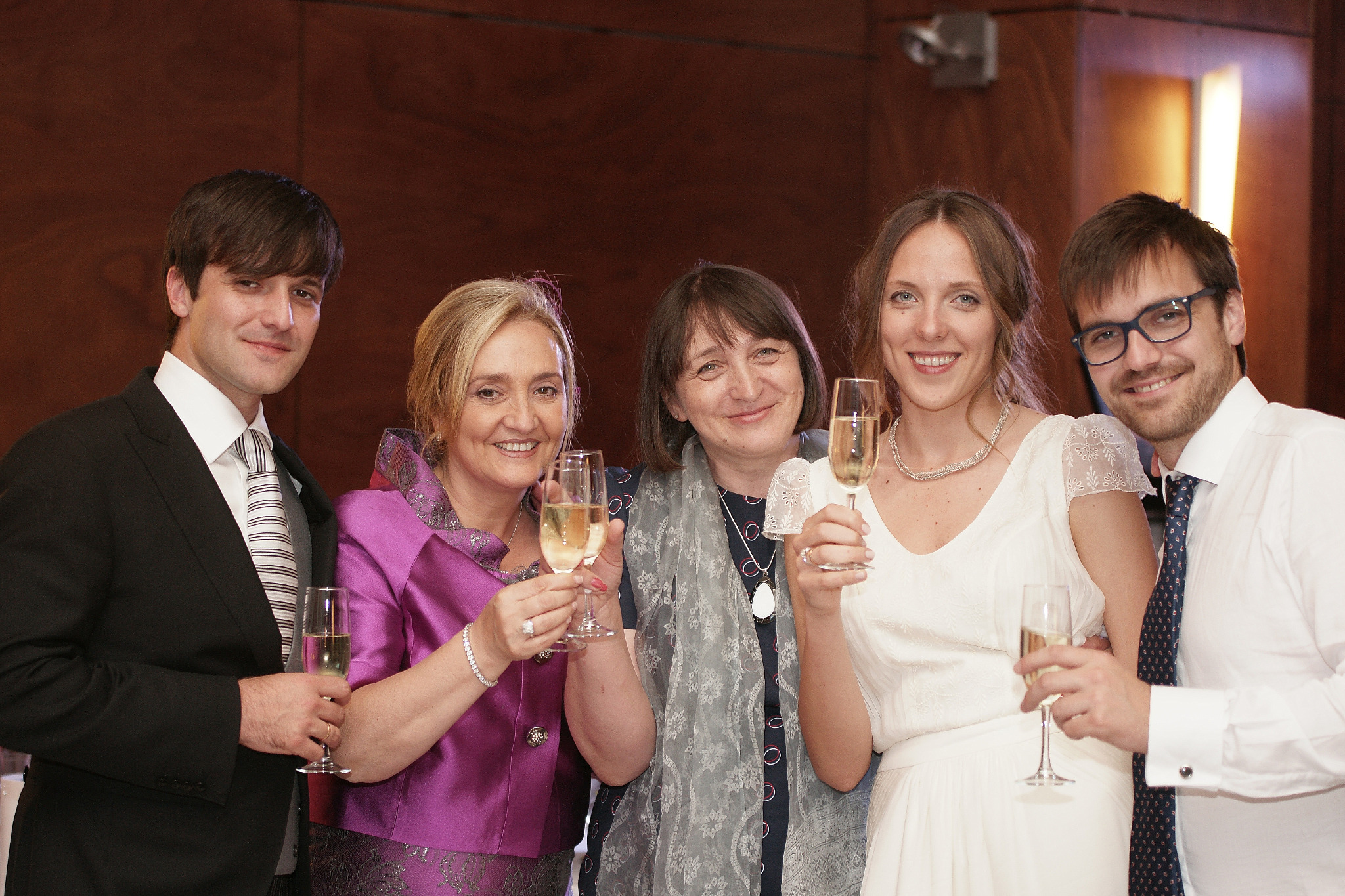 Grupa pięciu osób w eleganckich strojach wznosi toast szampanem na tle ciemnego drewna.