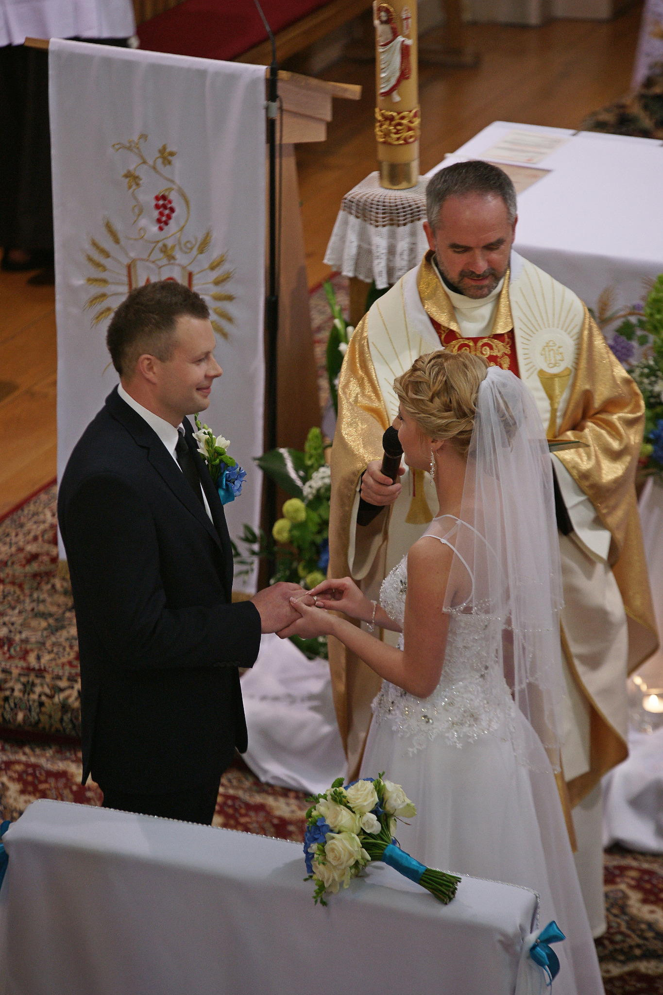 Para młoda wymienia obrączki podczas ceremonii ślubnej w kościele, w tle ksiądz w szatach liturgicznych i dekoracja z motywem winogron.
