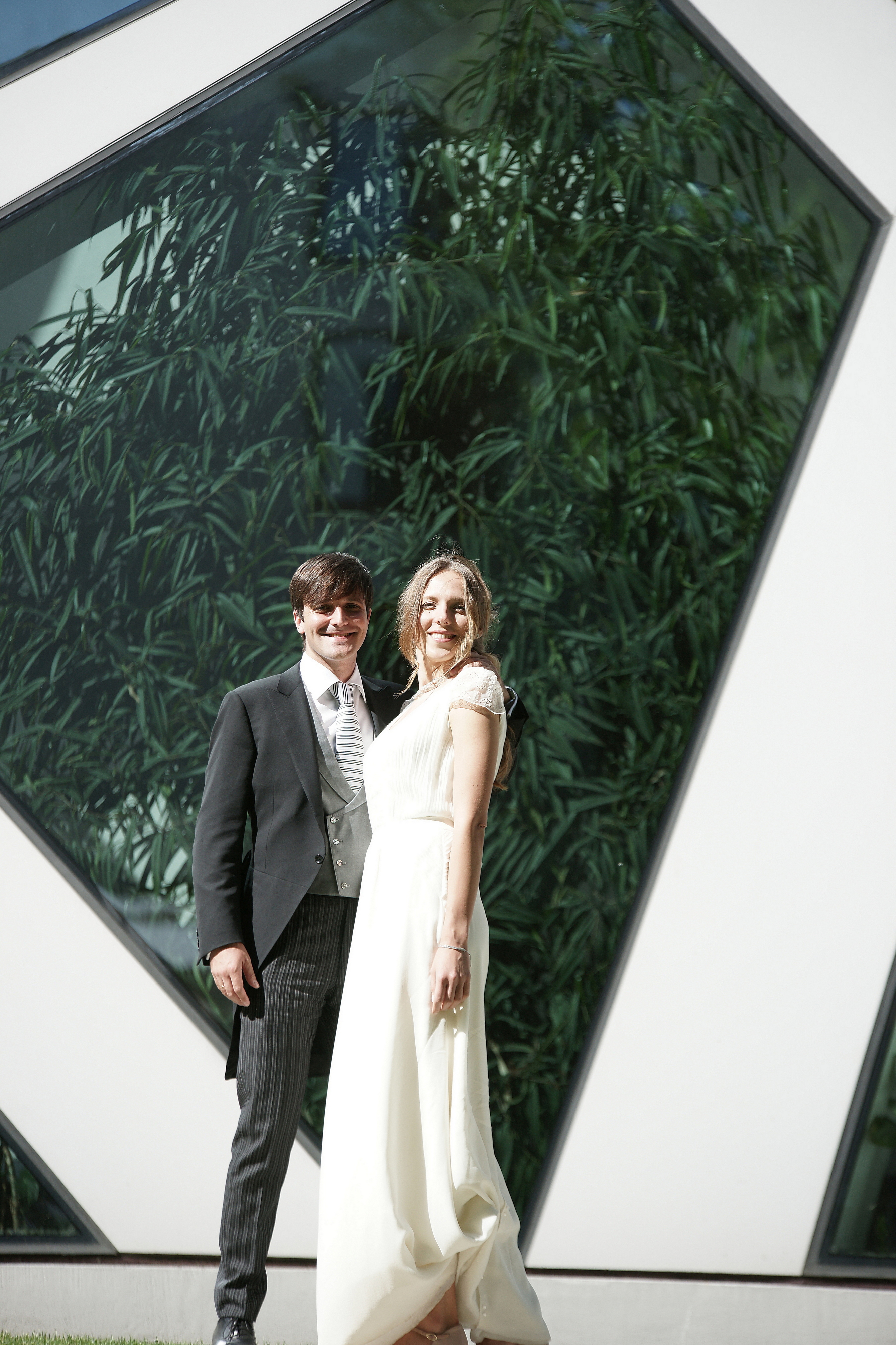 Para młoda w strojach ślubnych pozuje na tle nowoczesnej fasady budynku z geometrycznymi oknami, w których odbija się zieleń.