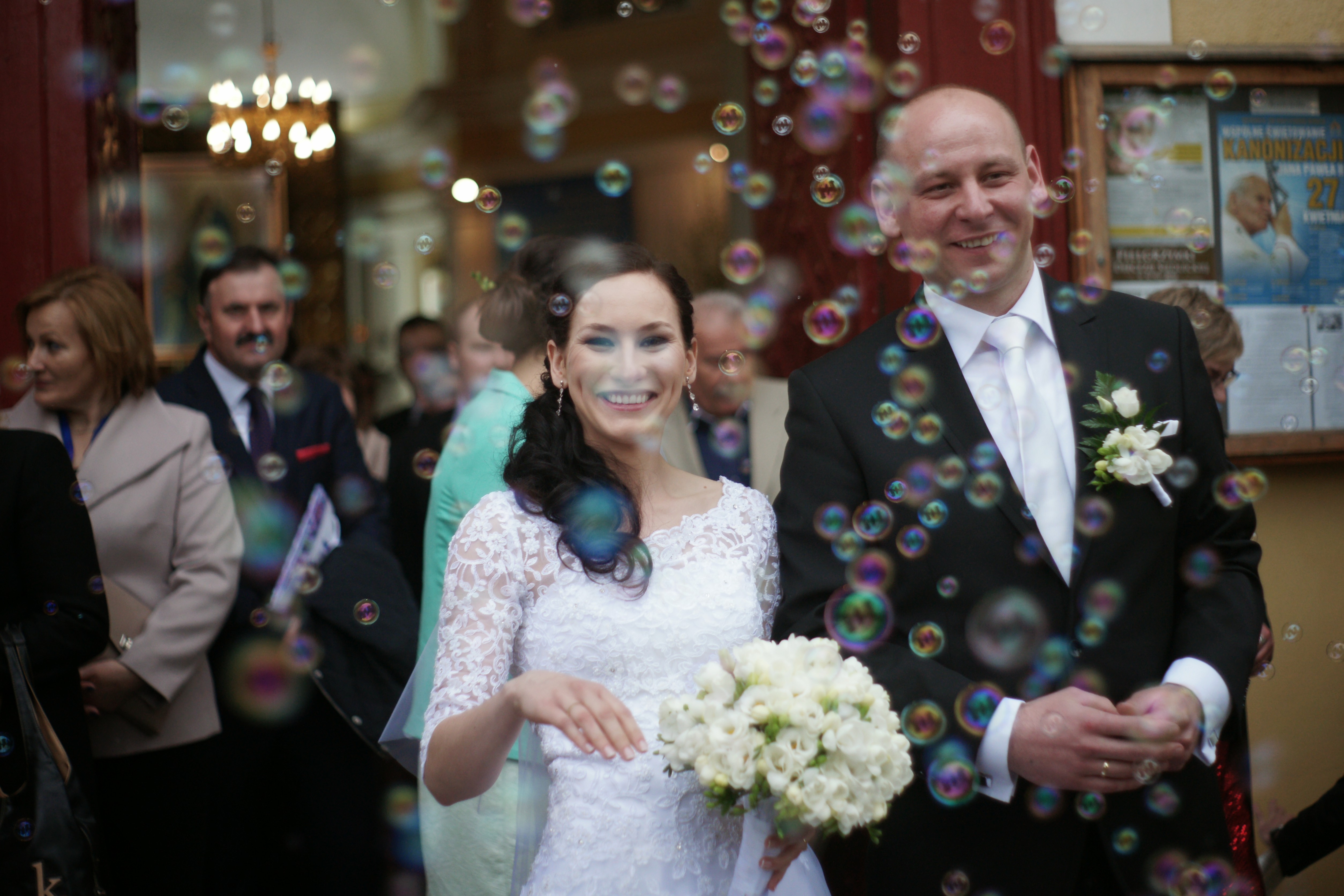 Uśmiechnięta para młoda wychodzi z kościoła, otoczona bańkami mydlanymi, panna młoda w koronkowej sukni trzyma bukiet białych kwiatów, pan młody w czarnym garniturze z butonierką.