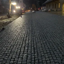 Nowo ułożona, ciemna nawierzchnia z kostki, rozświetlona reflektorami koparki w tle, z elementami przygotowania podłoża widocznymi po lewej stronie.