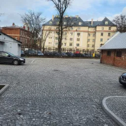 Obszerny parking wyłożony szarą kostką, z widocznymi samochodami w tle i budynkami o zróżnicowanej architekturze w tle.