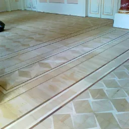 Wzór parkietu po renowacji w jasnych odcieniach drewna, z dekoracyjnymi bordiurami i geometrycznymi elementami, widoczne drzwi z ozdobnymi listwami i fragment kolumny w tle.